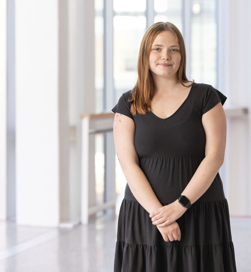A young woman wearing a black dress stands in the atrium of the RIT SHED