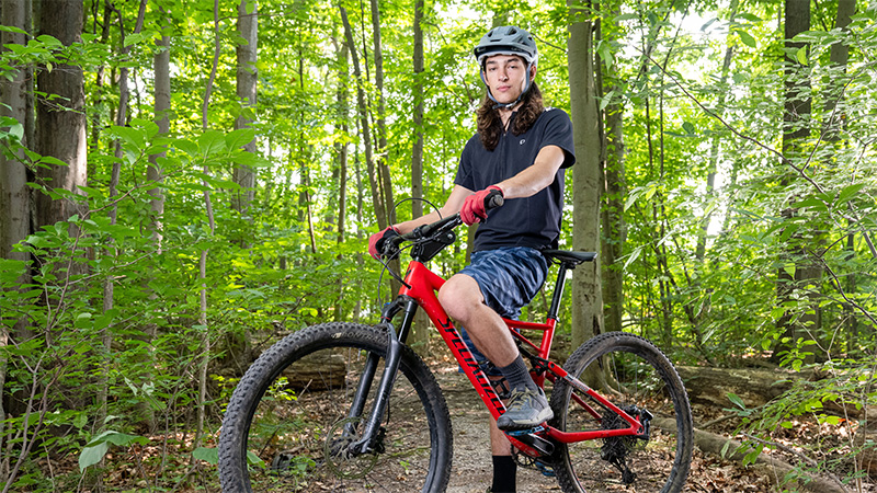 Student posing for a photo on his mountain bike, in a wooded trail.