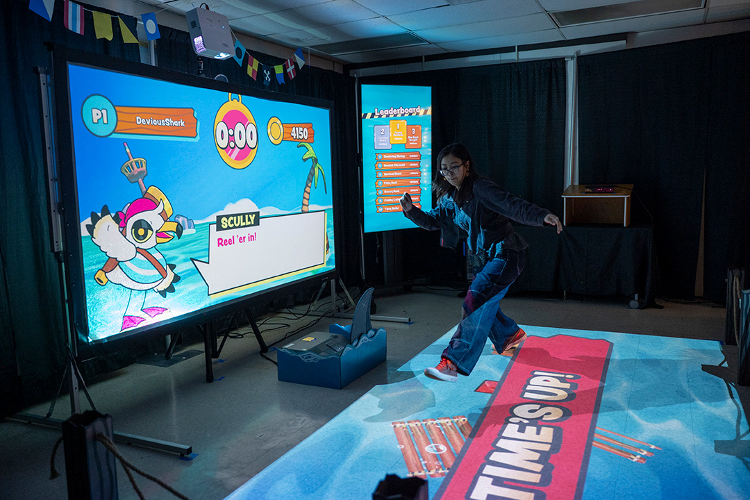 A seagull on a projected screen showing the Overboard game, with a water projected on to a floor. A student is in midair, jumping to land on wood boards to stay out of the projected water 