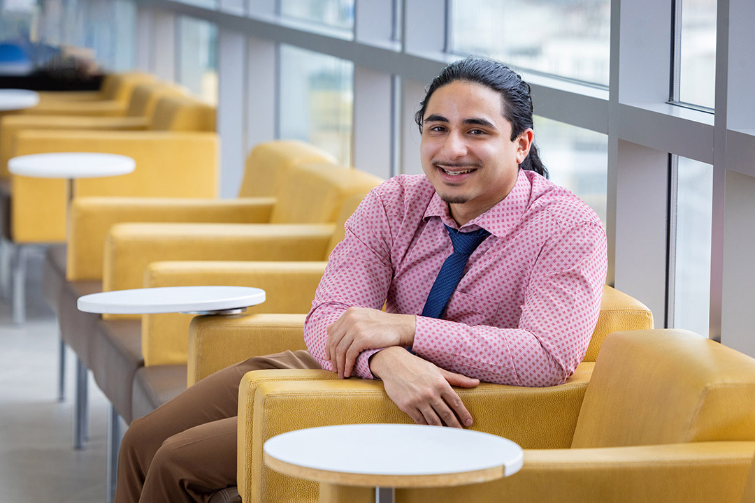 Gabe McMurtrie is wearing a pink patterned dress shirt and a navy tie and is seated in a modern lounge area with mustard-yellow armchairs and small white round tables.