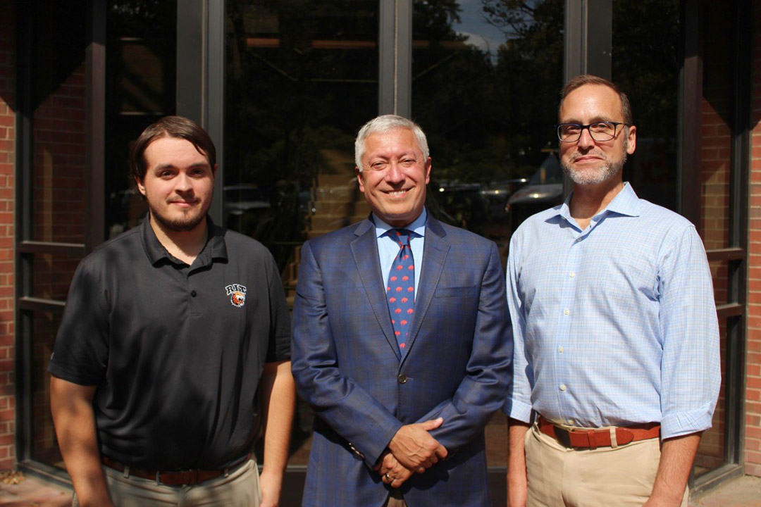 Three individuals stand in front of a glass entrance with brick framing. The person on the left is wearing a black polo shirt with a logo, the person in the center is dressed in a blue plaid suit with a patterned tie, and the person on the right is wearing a light blue button-down shirt with khaki pants and a brown belt.