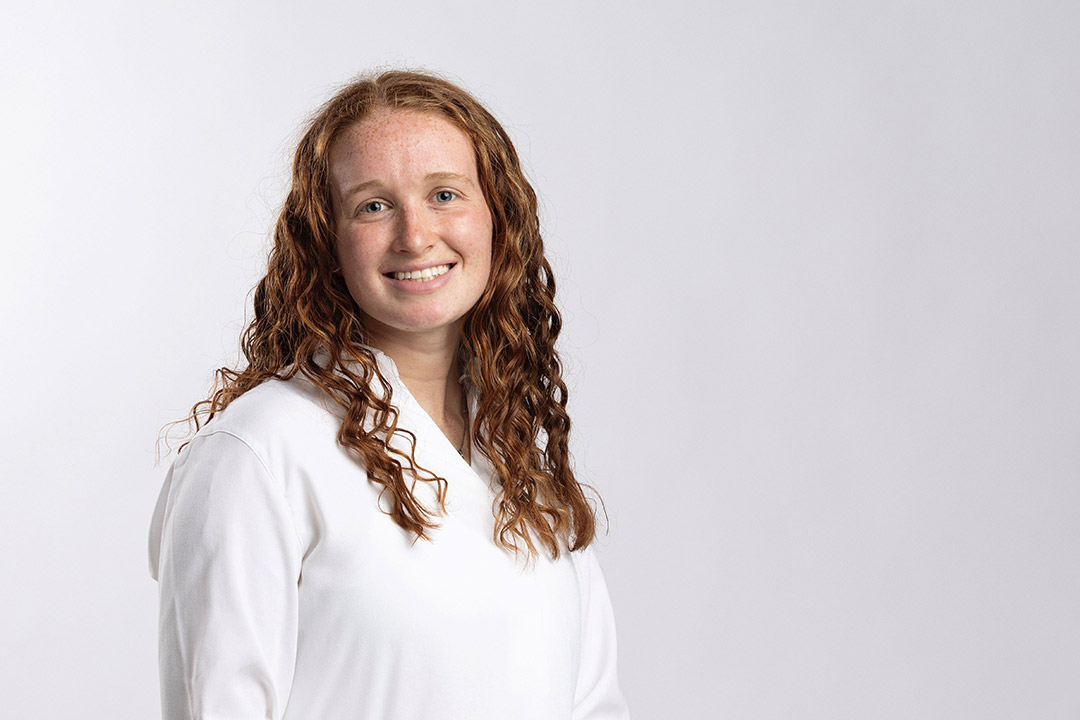 RIT student Madison Degenfelder has long, curly brown hair and is wearing a plain white top, posed against a neutral light gray background.