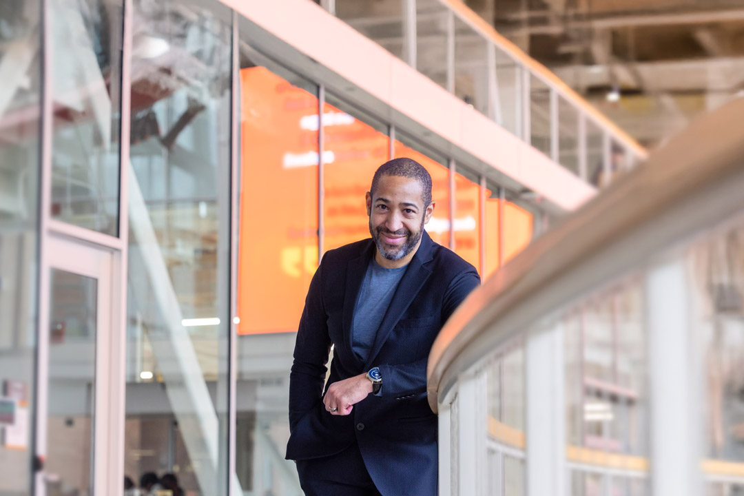 RIT alumn Jason Blythe is wearing a dark blazer and gray shirt. He is leaning casually on a railing inside a modern building with glass walls and an orange digital display in the background. 