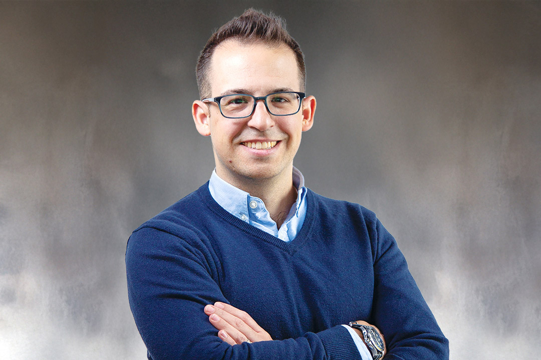 Ryan Larcome is wearing a dark blue sweater over a light blue collared shirt stands with arms crossed against a neutral gray studio background.