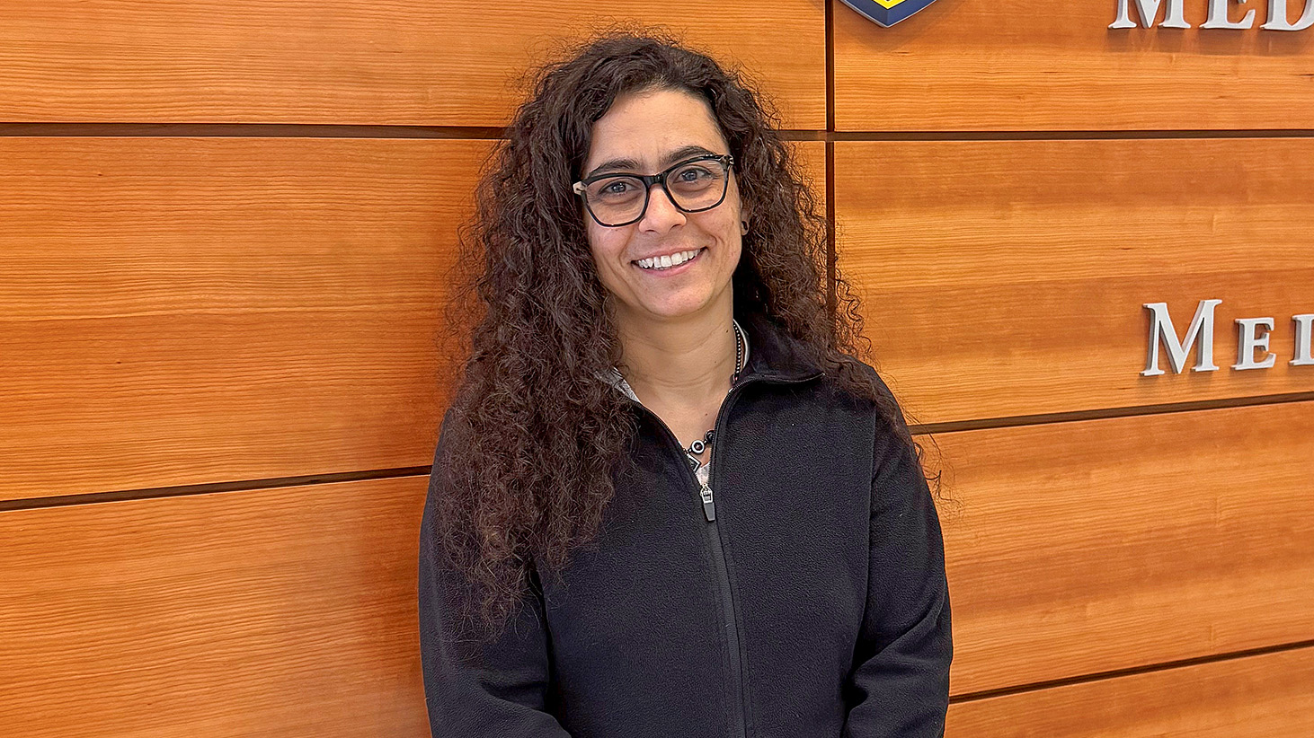 A smiling woman with long curly dark hair and black-rimmed glasses, wearing a black zip-up fleece jacket. She is standing indoors in front of a wall made of horizontal wooden panels.