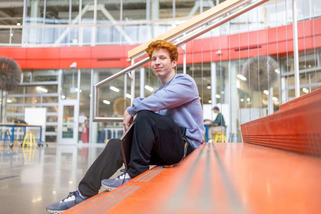 Benjamin Brundage in blue sweatshirt and black pants sits on orange stairwell in RIT SHED