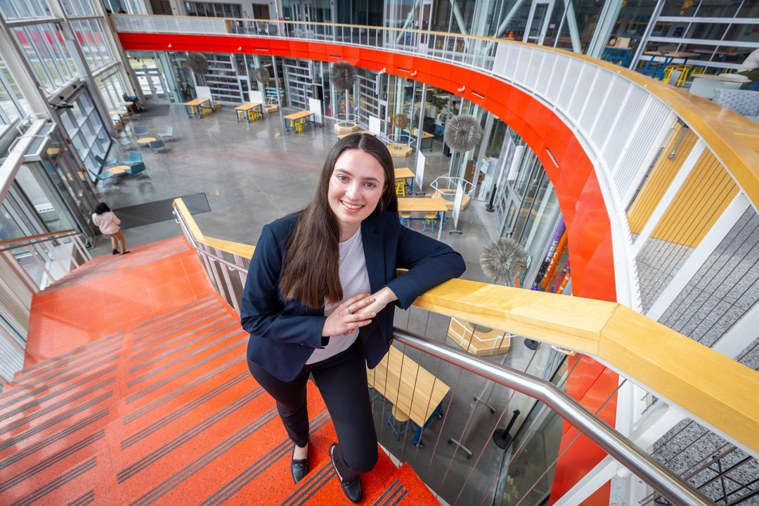 A young woman in business attire smiles at the camera while leaning on a railing at the top of a bright orange staircase inside a modern, multi-level building with glass walls, open seating areas, and a spacious atrium below.