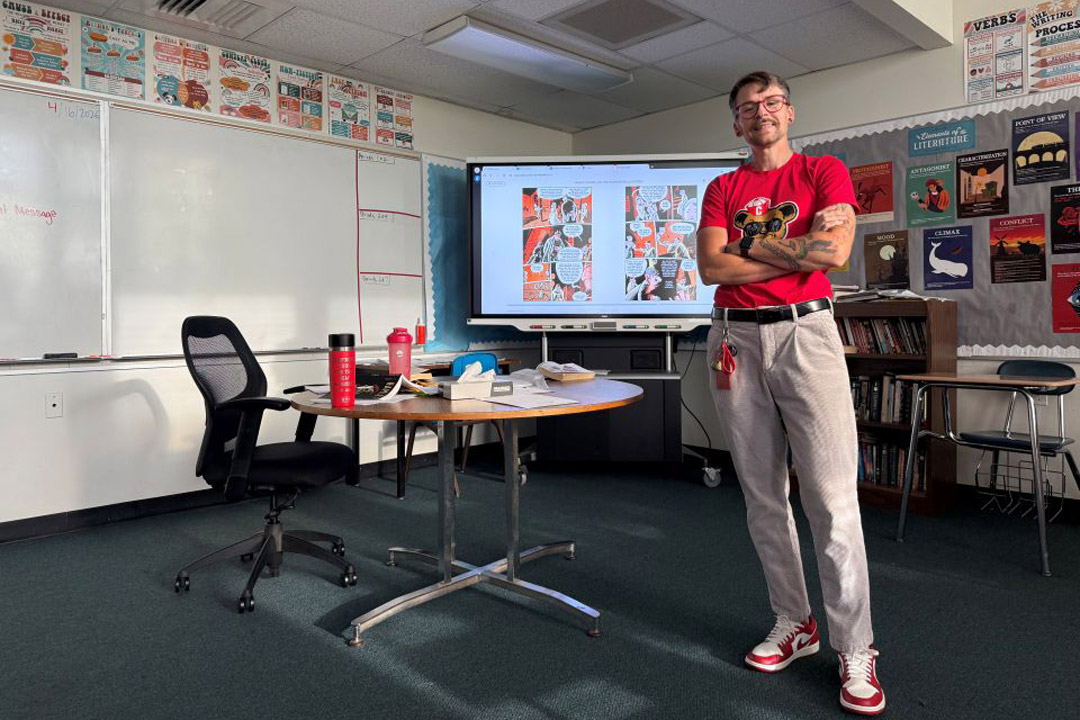 Blake Culley, an NTID graduate student, smiles while standing in a bright, modern classroom at RIT, wearing a "Be Kind" t-shirt and gesturing toward a whiteboard with educational notes.