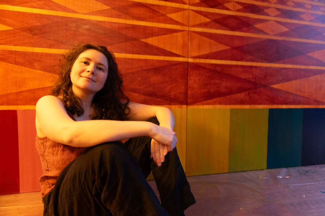 A woman with long curly brown hair sits on the floor, leaning forward with her arms crossed over her knees. She is smiling gently at the camera against a vibrant backdrop of a large wall painting featuring orange and red geometric diamond patterns above a row of multicolored vertical stripes.