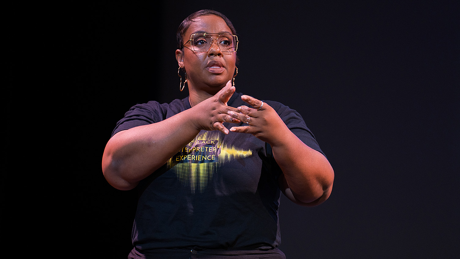 Yalanda Allgood standing on a dark stage, speaking and signing. She is wearing a black t-shirt that reads Black Interpreter Experience.