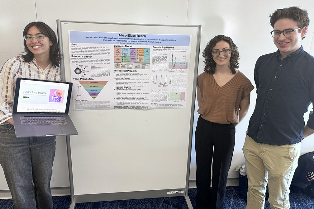 Three master's students. One standing to the left of a research poster holding a laptop. Two standing to the right of the research poster and smiling.
