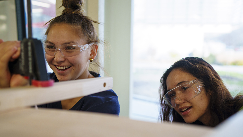 Two students smile while using woodworking tools.