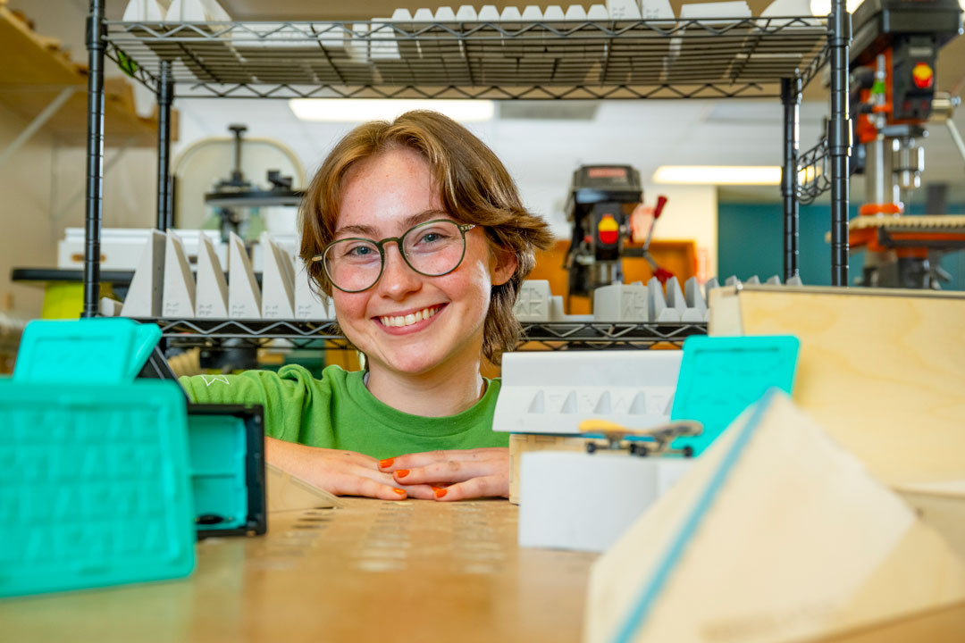 Person wearing glasses and a green shirt leans on a workbench in a makerspace or fabrication lab. In the foreground are miniature skateboarding ramps and 3D-printed objects, with shelving, tools, and workshop equipment visible in the background.