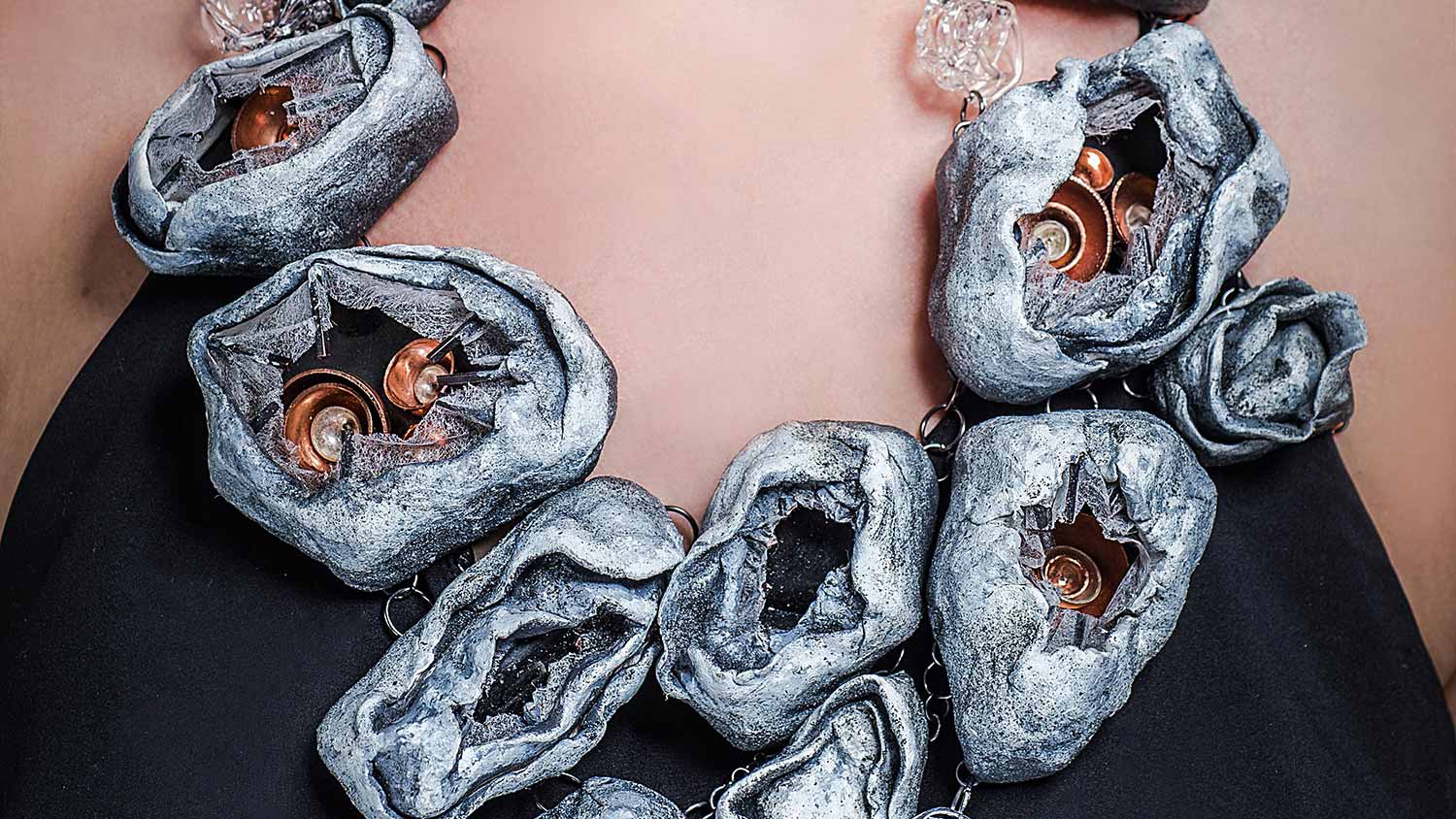 a necklace art piece with folded up gray pockets with small, concentric copper bowls topped with a diamond.