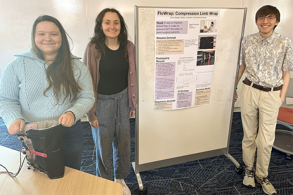 Three students standing on either side of a research poster on FloWrap and smiling.