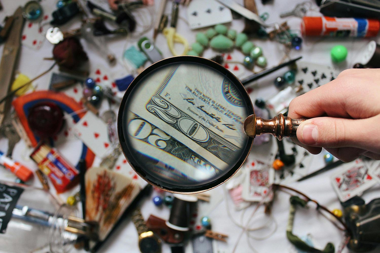 a cluttered desk with a person holding a magnifying glass to a U S 20 dollar bill.
