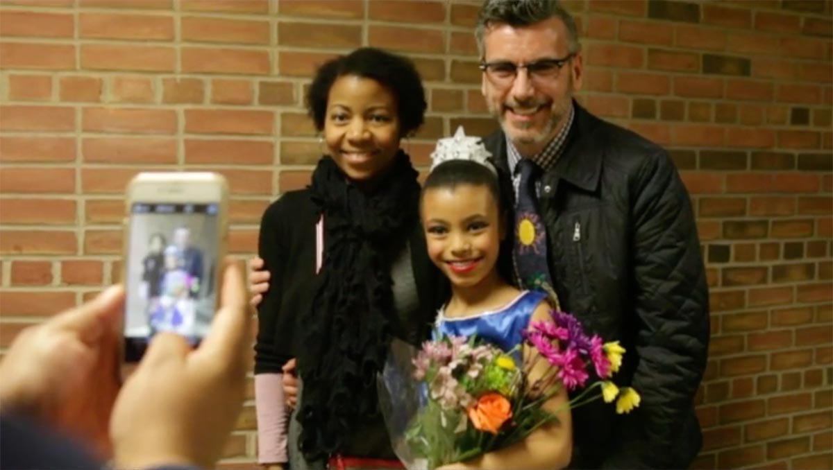 two adults and a child pose for a photo. The child is wearing a gown and tiara and holding a bouquet.