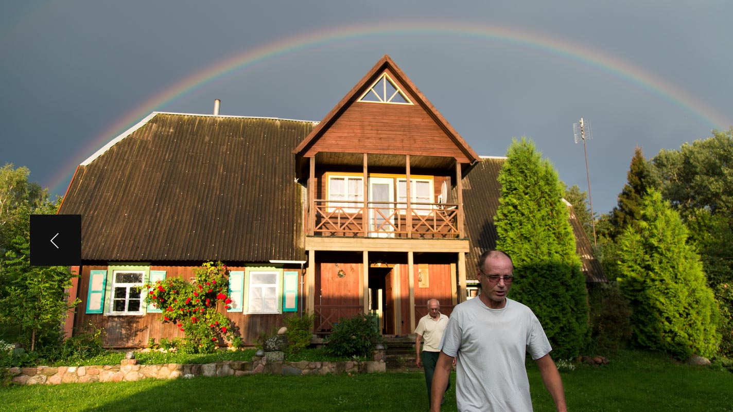 two people walking away from a farmhouse style building, with a large rainbow in the sky behind the house.
