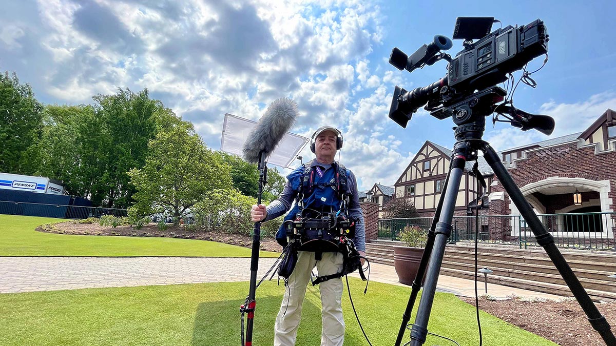 man standing outside a golf course club house with sound and video equipment.