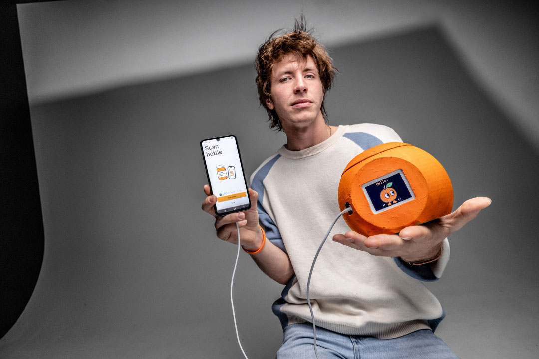 Student demonstrates a prototype consisting of a smartphone app and a connected orange, cylindrical device with a digital display. The smartphone screen shows a bottle-scanning interface, while the device displays a cartoon-style graphic. The student is seated in a studio setting with a neutral gray background, holding the phone in one hand and the prototype in the other.