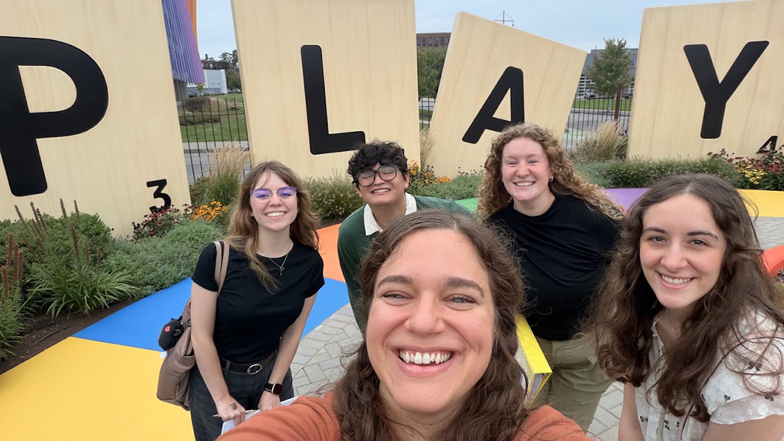 RIT MST students take a group selfie outside the Strong Museum of Play, with big scrabble letters behind them spelling Play.