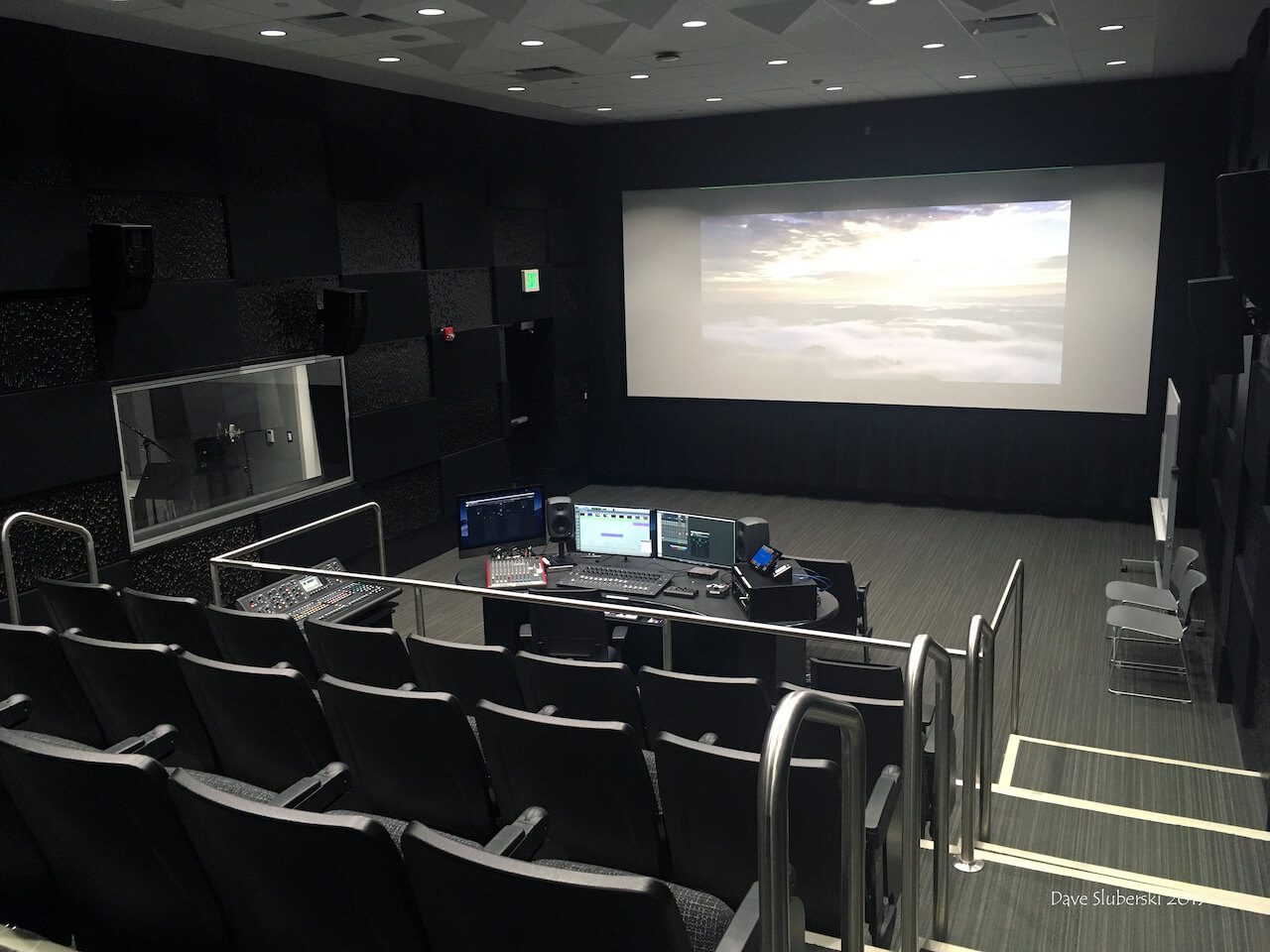 a small film screening room with auditorium seating, a large screen, and a work station with several monitors and sound mixing equipment.